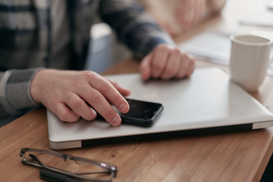 Hands reaching for a smartphone resting on a closed laptop at a wooden desk, a focus block cut short