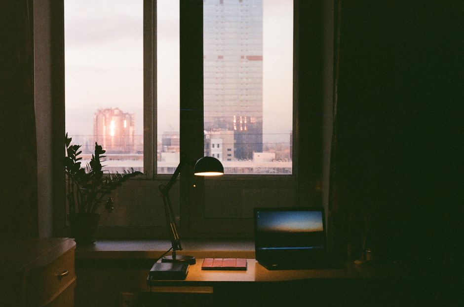 Desk with a laptop and a lit lamp by a window as the city goes dark at dusk